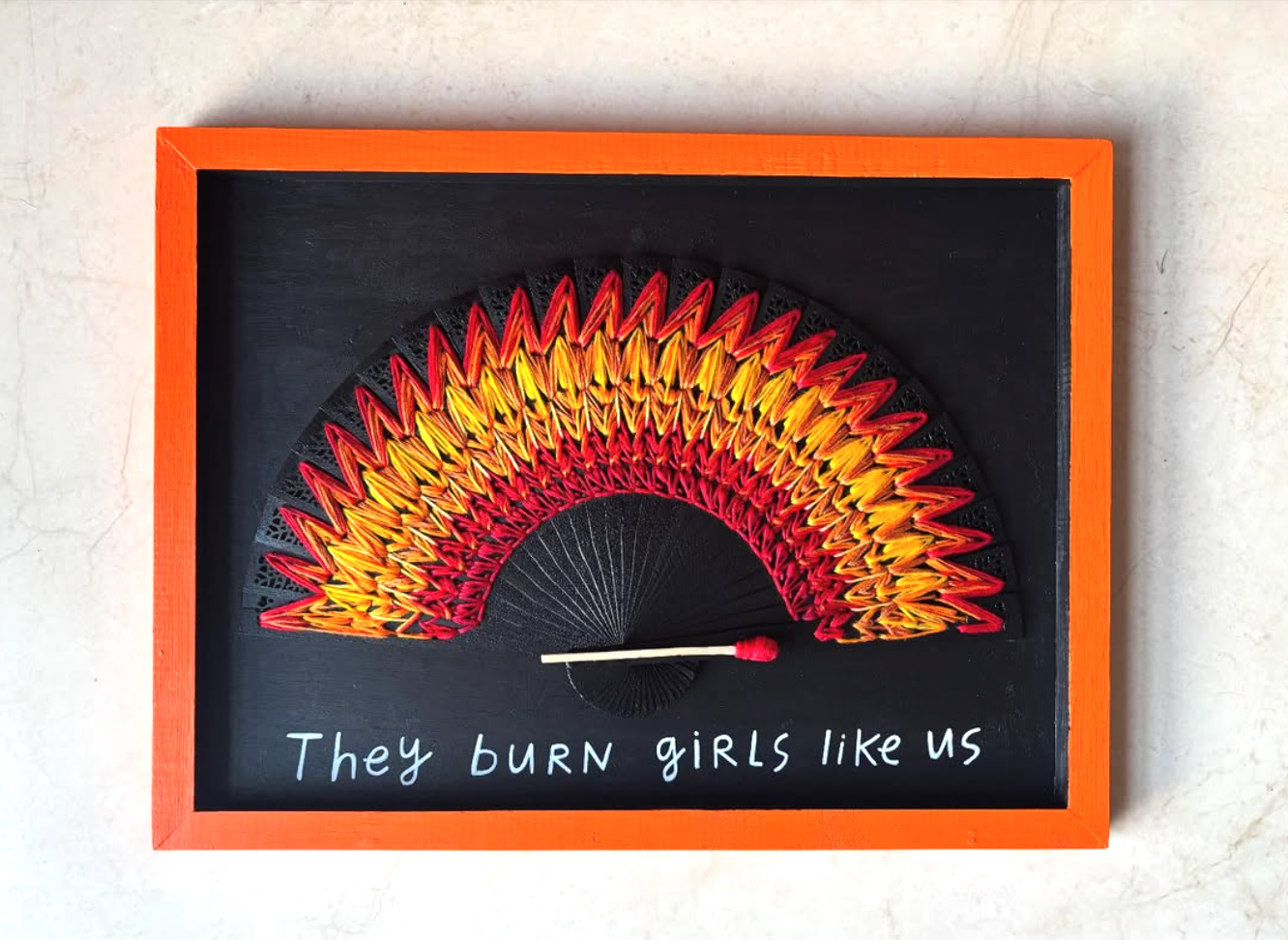 Framed artwork with a matchstick and colorful embroidered flame fan on a black background. Hand lettering.