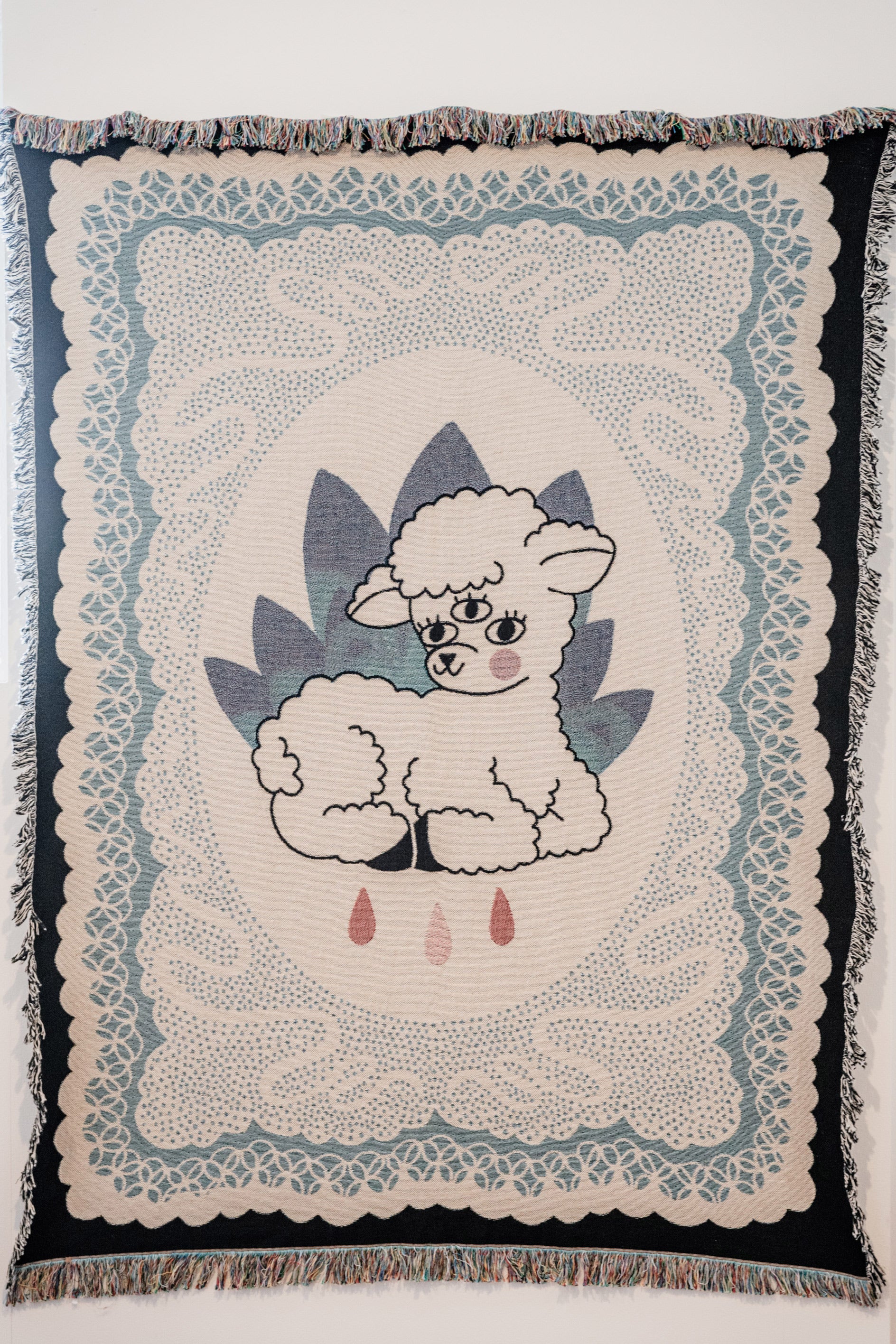 A decorative tapestry featuring a central lamb design with a blue and white color palette, surrounded by a detailed floral pattern, and fringed with a multicolored border.