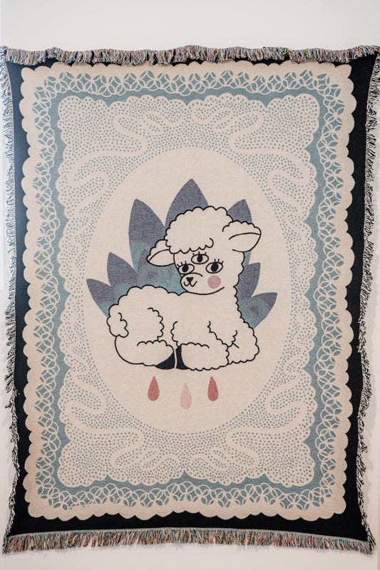 A decorative tapestry featuring a central lamb design with a blue and white color palette, surrounded by a detailed floral pattern, and fringed with a multicolored border.